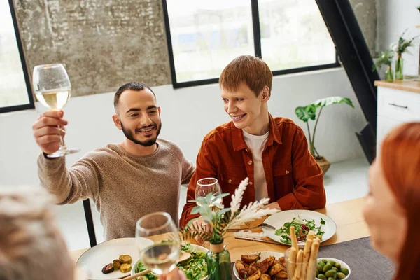 Bearded man toasting with wine near happy gay partner and delicious family dinner in modern kitchen — Stock Photo