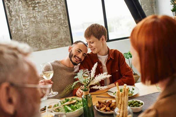 Overjoyed gay man with wine glass leaning on redhead boyfriend during supper with blurred parents — Stock Photo