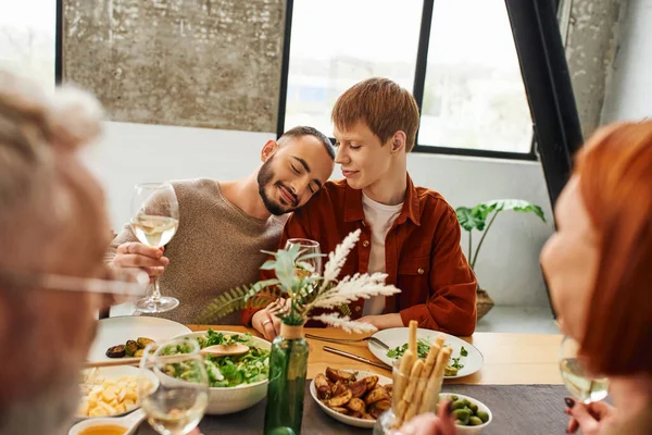 Pleased gay man with wine glass leaning on redhead boyfriend near blurred parents and delicious supper in kitchen — Stock Photo