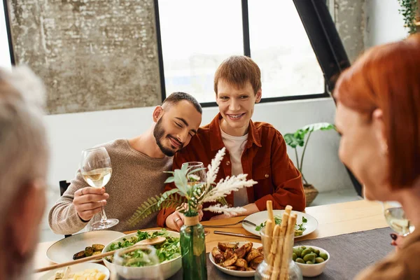 Joyful bearded man with glass of wine leaning on happy gay partner during family dinner in kitchen — Stock Photo