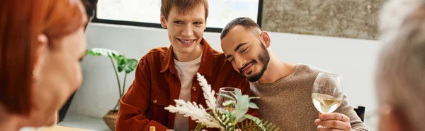 Rousse homme regardant les parents flous près heureux partenaire gay et verres à vin à la maison, bannière — Photo de stock