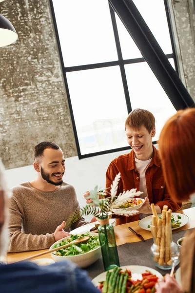 Redhead gay man holding salad near boyfriend during family supper with blurred parents in modern kitchen — Stock Photo