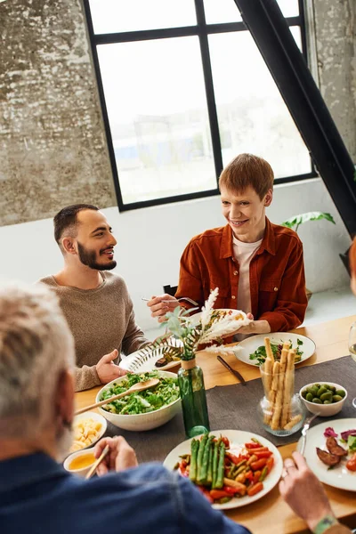 Pelirroja mirando a los padres mientras sirve ensalada cerca de la pareja gay durante la cena familiar - foto de stock