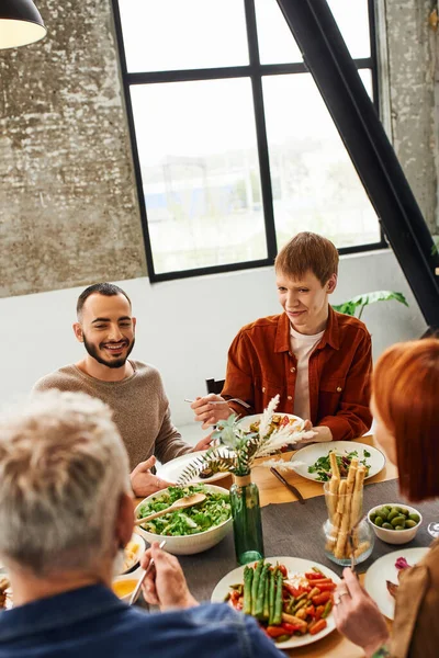 Рыжий гей улыбается рядом с бородатым парнем и размытыми родителями с вкусной едой, подаваемой на кухне — стоковое фото