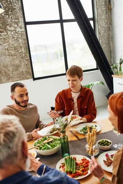 Бородатый гей разговаривает с рыжим парнем и размытыми родителями во время семейного ужина на кухне — стоковое фото
