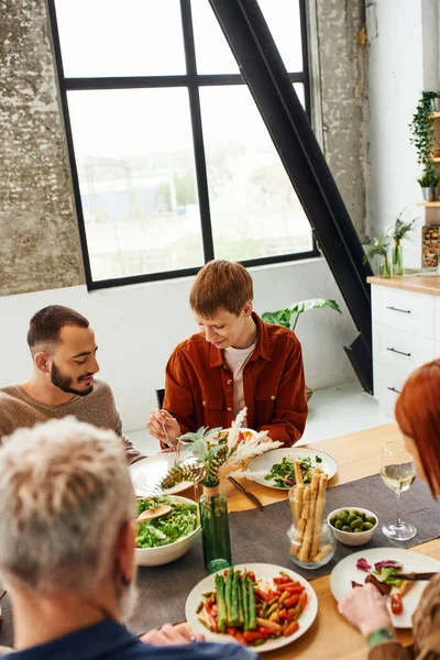 Высокий угол зрения рыжеволосый мужчина, имеющий семейный ужин с гей-партнером и размытые родители на современной кухне — стоковое фото