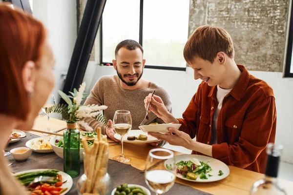 Веселый рыжий мужчина, подающий еду рядом с гей-партнером и размытая мать во время семейного ужина на кухне — стоковое фото