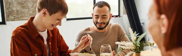 Redhead man serving meal near happy boyfriend and blurred mother in kitchen, banner — Stock Photo
