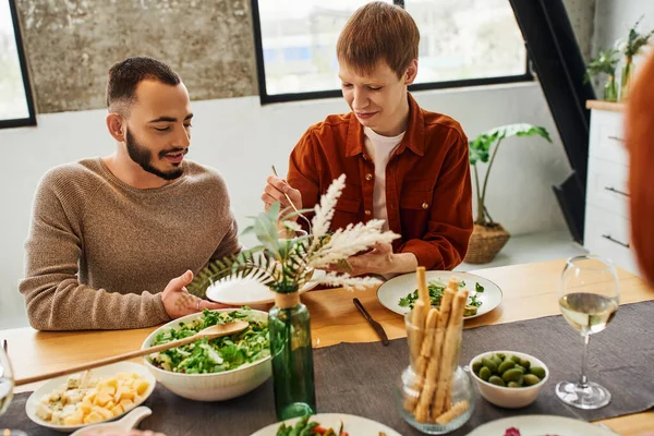 Щасливий гей чоловік подає їжу біля хлопця біля смачної вечері на сучасній кухні — стокове фото