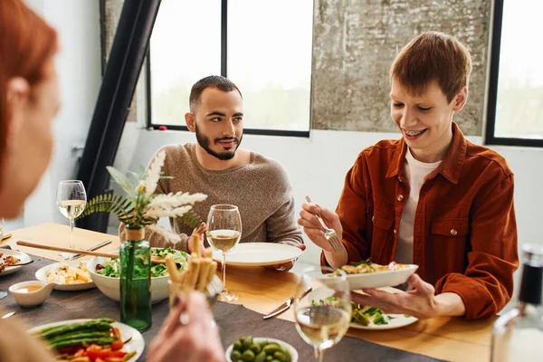 Barbudo hombre mirando feliz gay pareja servir comida durante familia cena en moderno cocina - foto de stock
