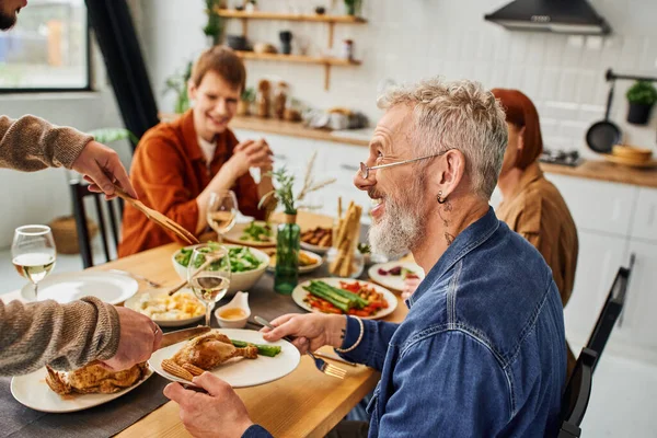 Gay man cutting delicious chicken near happy father during family supper in kitchen — Stock Photo