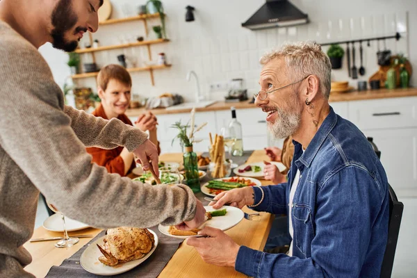 Smiling bearded man looking at gay man serving chicken during supper with family in kitchen — Stock Photo