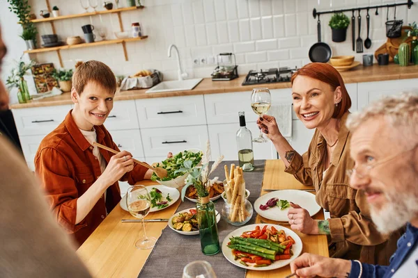 Щасливий гей чоловік з батьками дивиться на розмитого хлопця, маючи смачну вечерю на кухні — стокове фото