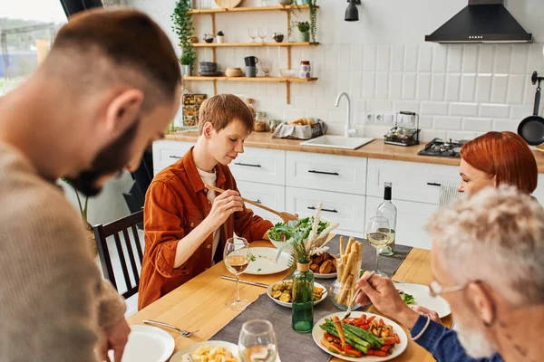 Pelirroja gay hombre celebración ensalada mientras tener cena con familia y borrosa novio en cocina - foto de stock