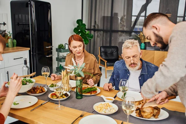 Bearded gay man cutting grilled chicken during family dinner with boyfriend and parents in kitchen — Stock Photo