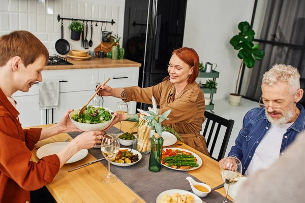 Mulher ruiva feliz servindo salada durante deliciosa ceia com marido e filho na cozinha moderna — Fotografia de Stock