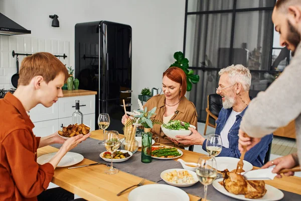 Desenfocado gay hombre corte a la parrilla pollo cerca de familia servir delicioso comida durante familia cena - foto de stock