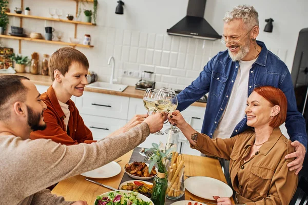 Joven gay hombre tintineo vino vasos con novio y feliz padres durante familia cena en cocina - foto de stock