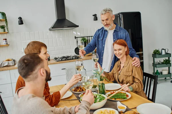 Bearded man looking at young gay couple and toasting with wine glass near delicious supper in kitchen — Stock Photo