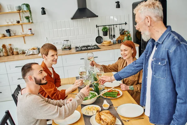 Joven pelirrojo hombre tintineo copas de vino con novio y padres cerca de la cena servido en la cocina moderna - foto de stock