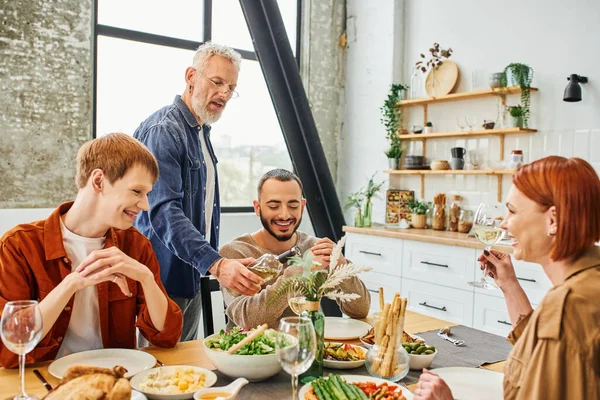 Bearded man showing wine bottle to cheerful gay couple during family supper in modern kitchen — Stock Photo