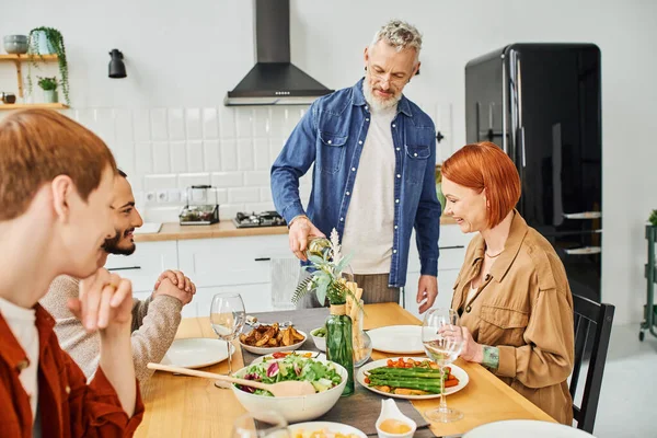 Bärtiger Mann schenkt Wein in der Nähe einer glücklichen Familie ein, die in der modernen Küche leckeres Abendessen einnimmt — Stockfoto