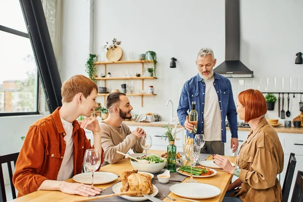 Smiling man standing with wine bottle near family and tasty supper served in kitchen — Stock Photo