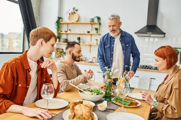 Barbudo hombre de pie con botella de vino cerca de hijo con gay pareja durante la cena familiar en la cocina - foto de stock