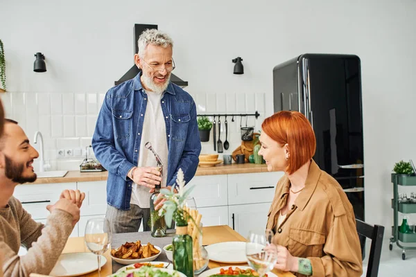 Весёлый бородатый мужчина открывает бутылку вина во время ужина с женой и сыном на кухне — стоковое фото