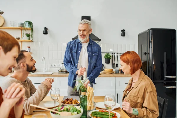 Sonriente barbudo hombre apertura vino botella cerca familia cena con hijo y gay hombre en cocina - foto de stock
