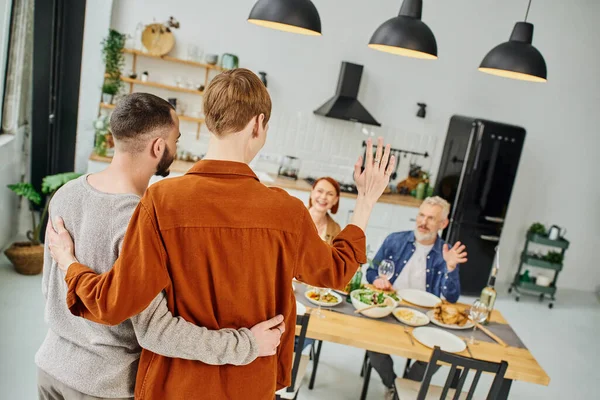 Рыжий гей обнимает парня и машет рукой родителям, ужинающим на кухне — стоковое фото