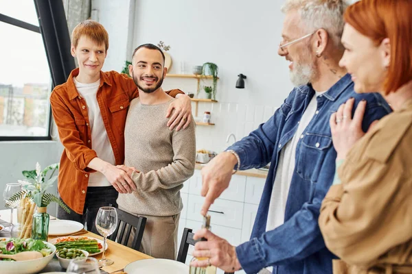 Happy gay couple embracing near blurred parents and delicious supper in kitchen — Stock Photo