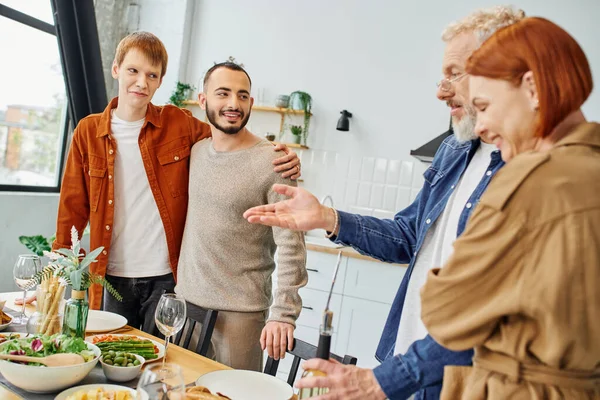 Бородатый мужчина указывает на вкусный семейный ужин возле счастливой гей-пары на кухне — стоковое фото