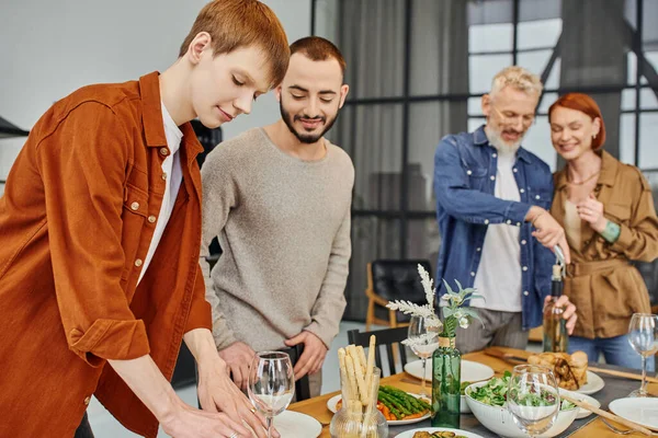 Happy gay couple setting family supper near parents on blurred background in kitchen — Stock Photo