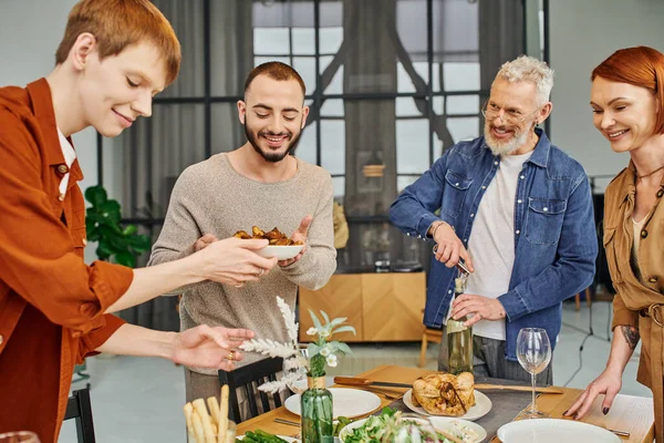 Cheerful gay couple serving grilled vegetables near father opening wine in kitchen — Stock Photo