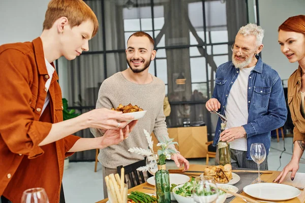Redhead gay man holding grilled vegetables near boyfriend and happy parents in kitchen — Stock Photo