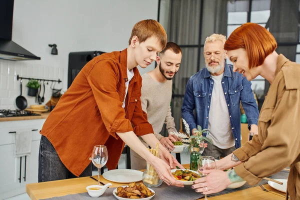Pelirroja gay hombre con madre servir a la parrilla verduras en mesa con deliciosa cena en cocina - foto de stock
