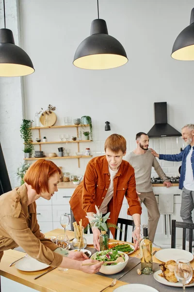 Young gay man setting table with redhead mother for family supper in kitchen — Stock Photo