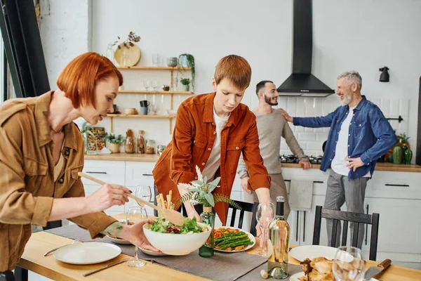 Redhead woman mixing salad near son setting family supper on table in kitchen — Stock Photo