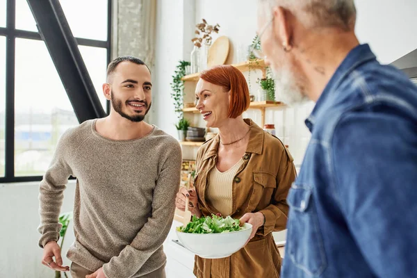 Happy redhead woman holding bowl with fresh salad near son and blurred husband in kitchen — Stock Photo