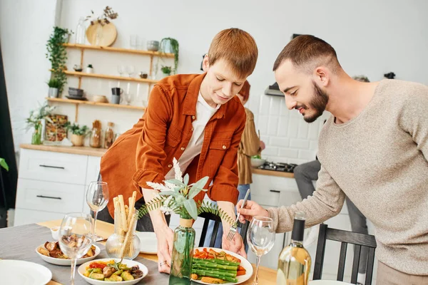 Gay man holding fork near boyfriend holding grilled asparagus near table with family supper in kitchen — Stock Photo