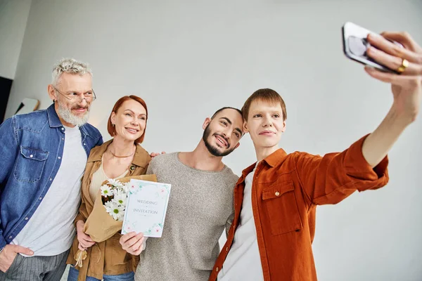 Feliz gay hombre tomando selfie con novio y padres holding flores y boda invitación tarjeta - foto de stock