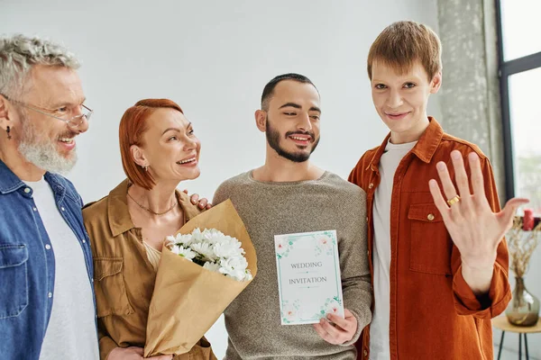 Fröhlicher schwuler Mann zeigt Ehering neben Freund mit Einladungskarte und glückliche Eltern mit Blumen — Stockfoto