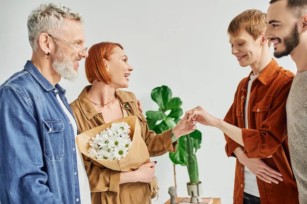 Joven gay hombre mostrando boda anillo a feliz madre cerca novio y padre en casa - foto de stock
