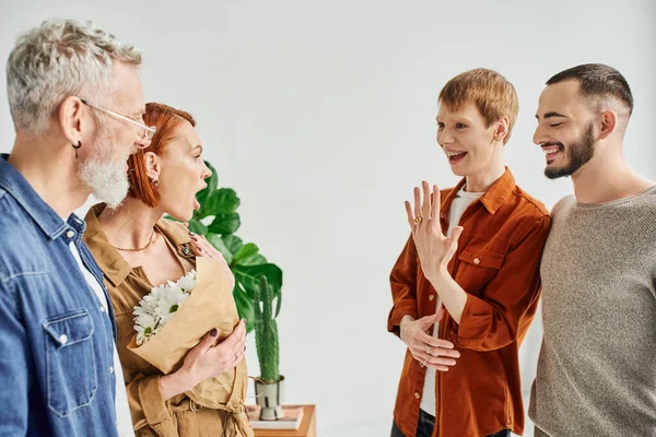 Hombre gay lleno de alegría mostrando anillo de boda a los padres sorprendidos en casa - foto de stock