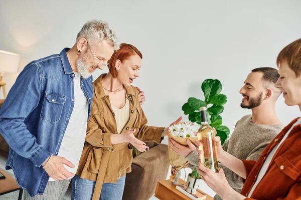 Young gay couple visiting happy parents at home while holding flowers and bottle of wine — Stock Photo