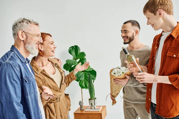 Excited woman showing wow gesture near son with gay partner holding flowers and wine at home — Stock Photo