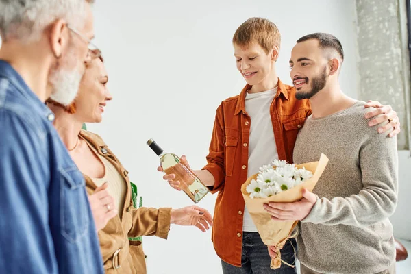 Веселая гей-пара с цветами и бутылкой вина возле родителей дома — стоковое фото