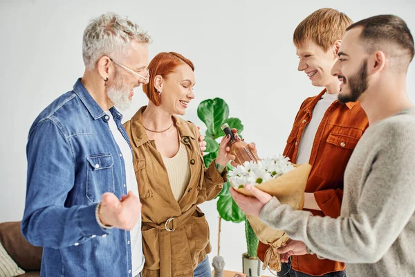 Fröhliches homosexuelles Paar schenkt Blumen und Weinflasche, während es glückliche Eltern zu Hause besucht — Stockfoto