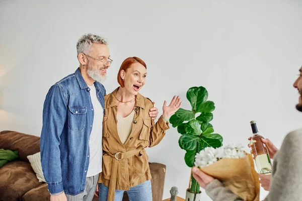 Amazed woman showing wow gesture near gay couple with flowers and wine bottle at home — Stock Photo